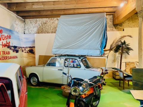 Museumsaustellung zeigt einen Trabant mit Dachzelt, im Vordergund eine orangene Kreidler mit verchromten Tank, im Vordergrund des linken Bildrandes ein schmaler Teil eines weiteren Autos mit weißem Dach und roter Karrose